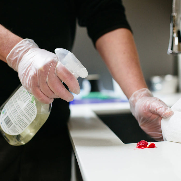 Clear Industrial Vinyl Gloves on Hand Wiping Red Substance Off Counter with Paper Towel and Cleaning Solution 